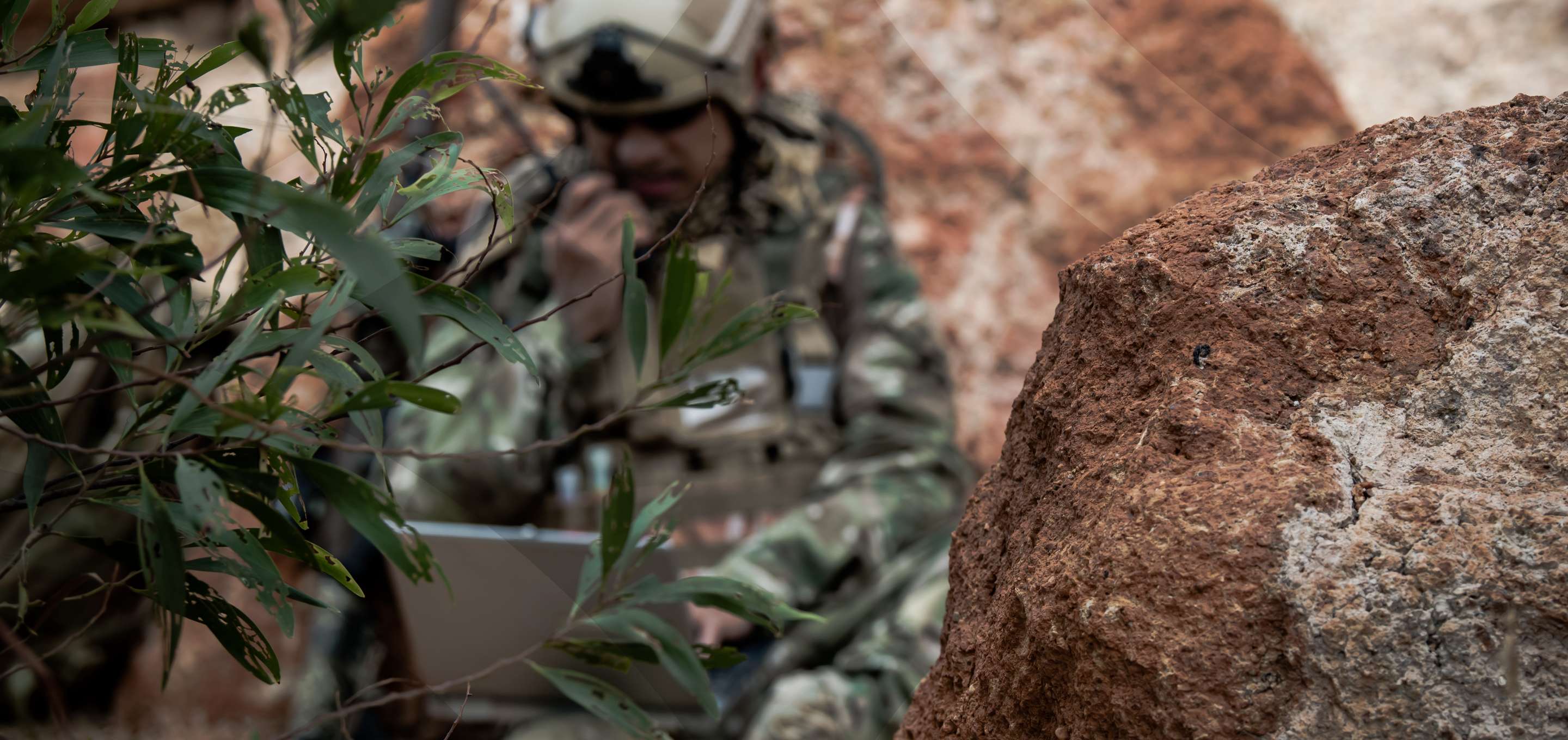 a soldier hidden between rocks and bushes using a rugged laptop